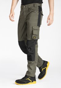 Pantalons cargo vert olive avec des accents noirs, comportant de multiples poches, des genoux renforcés et des coutures jaunes. Associés à des chaussures noires et jaunes.