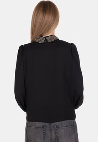 Blusa nera con maniche a sbuffo e colletto adornato di perline. Tessuto liscio, chiusura con bottone sul retro, abbinata a pantaloni in denim grigi.