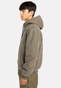 Hoodie vert olive avec grande poche kangourou, capuche réglable à cordon de serrage et poignets côtelés. Fabriqué en tissu doux et texturé. Vue de profil.