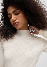 Kremfarget ribbestrikket turtleneck-genser med strukturert stoff, med et logomerke på venstre bryst og en tettsittende design.