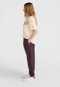 Beige katoenen t-shirt met zwarte tekst, gecombineerd met paarse sweatpants met dezelfde tekst, gedragen met witte sneakers. Zijaanzicht.