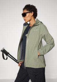 Light green waterproof jacket with a hood, zip closure, elastic cuffs, and two side pockets. Paired with black pants and a hiking pole.