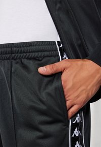 Main dans la poche d'un pantalon de sport noir avec ceinture élastique et bande blanche avec logo sur le côté, porté avec une veste noire et une chemise blanche.