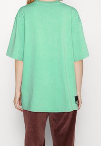 Osoba oblečená v volné světle zelené košili s krátkým rukávem a hnědých kalhotách, zachycena zezadu na jednobarevném pozadí.