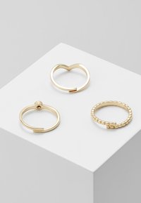 Tre anelli d'oro su un piedistallo bianco: uno a forma di cuore, uno con una fascia liscia e un'apertura, e uno con un design a perline.