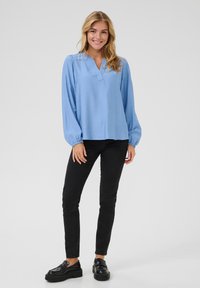 Blusa azzurro chiaro con scollatura a V e maniche lunghe a sbuffo, realizzata in tessuto morbido; abbinata a pantaloni neri aderenti e mocassini neri voluminosi.