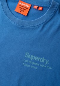 Blå bomulds t-shirt med rund halsudskæring, der har ordet "Superdry" og bynavne trykt i lys grøn på forsiden. Plejelabel inkluderet.