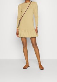 Béžové šaty s dlouhým rukávem a volánem na spodním okraji, vyrobené z měkké látky. Doplnené hnědými sandály na šněrování. Minimalistický design a vypasovaný střih.