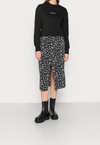 Sudadera negra corta con logo blanco, combinada con una falda negra a la altura de la rodilla que presenta un patrón floral blanco y una abertura frontal, llevada con botas negras.