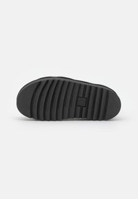 Semelle de chaussure noire en caoutchouc texturé, présentant des rainures horizontales, un bout rond et une empreinte de logo au centre.