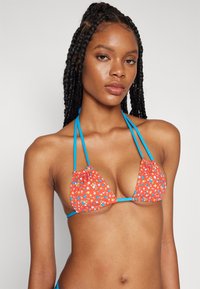Top bikini triangolare in tessuto rosso brillante con piccoli motivi floreali. Spalline blu creano un collo a halter e si legano sul retro. Texture liscia.