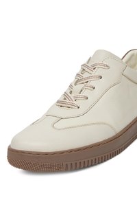 Kremowe skórzane sneakersy o zaokrąglonym kształcie, kontrastującej brązowej gumowej podeszwie, prążkowanych sznurówkach i teksturowanym wykończeniu po bokach.