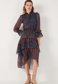 Chiffon-mekko, jossa on sininen ja ruskearaitainen paisley-kuvio, röyhelöhimo, korkea kaula-aukko ja pitkät hihat. Beige nilkkurit täydentävät asun.