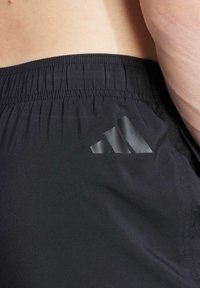 Bermudas deportivas negras con una cinturilla elástica, que cuentan con un logo de tres rayas en negro mate en la parte inferior izquierda. Textura suave.