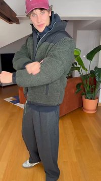 Green fleece jacket with a black nylon yoke, thick texture, paired with gray loose-fitting pants and a maroon cap. Indoor setting with plants.