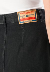 Nærbilde av svarte denimjeans med et lærmerke fra Diesel Industry som viser merke, kvalitet og årstall på linningen.