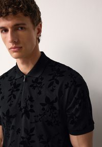 Rövid göndör hajú fiatal férfi, fekete, finoman virágmintás galléros pólóban, egy egyszerű fehér fal előtt áll.