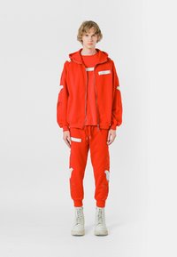 Sudadera con capucha roja con detalles bordados blancos, combinada con pantalones rojos a juego que presentan detalles similares. Usada con botas de color claro.