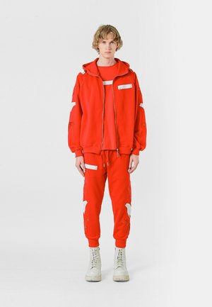 Sudadera con capucha roja con detalles bordados blancos, combinada con pantalones rojos a juego que presentan detalles similares. Usada con botas de color claro.