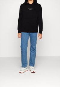 Felpa nera con cappuccio, dotata di tasca frontale e dettagli testuali, abbinata a jeans dritti di colore azzurro chiaro e sneakers bianche con dettagli rossi.