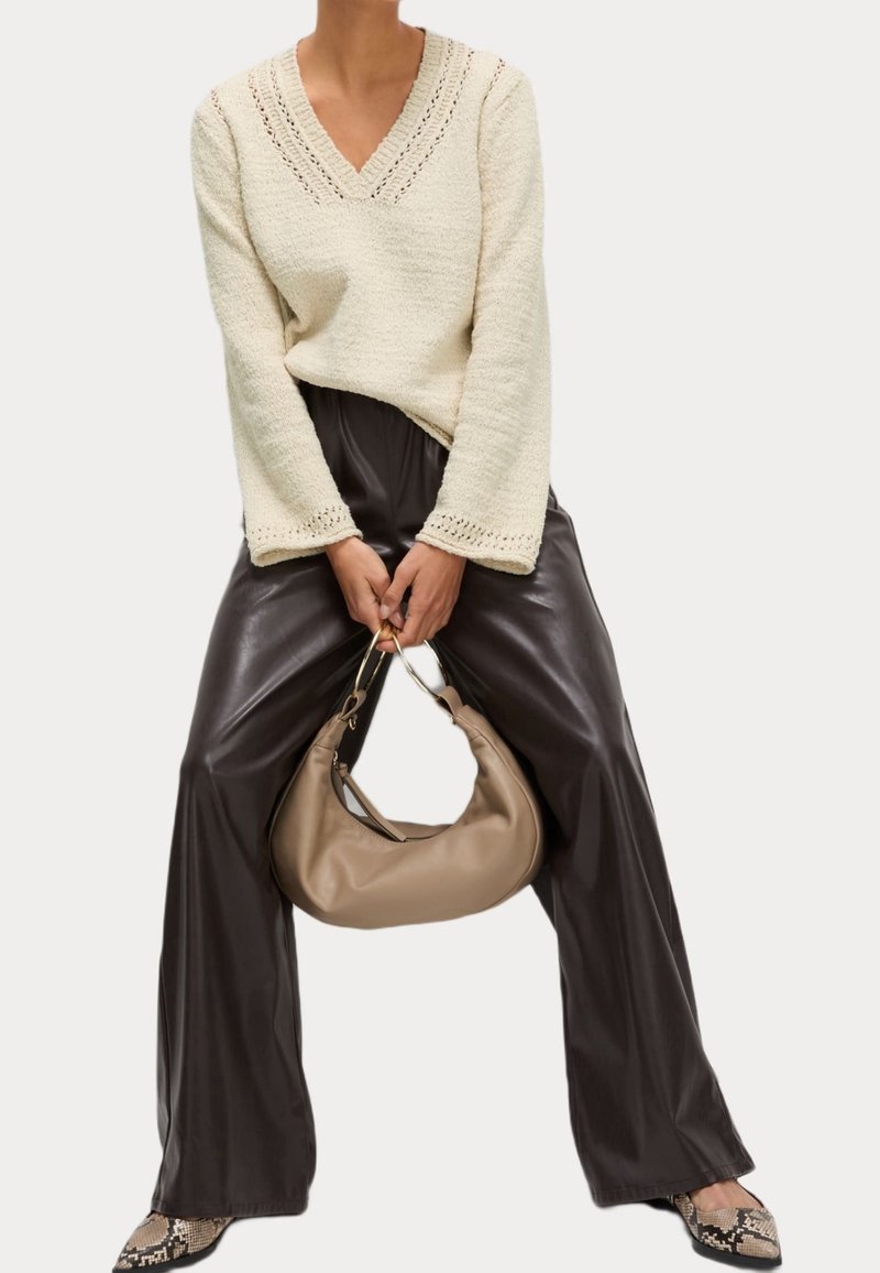 Femme portant un pull en maille crème, un pantalon en cuir marron foncé et des chaussures en peau de serpent, tenant un sac à main beige avec des anneaux dorés.