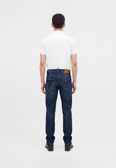 Homme debout, dos tourné, portant une chemise blanche à manches courtes, un jean bleu foncé et des chaussures noires sur un fond clair uni.