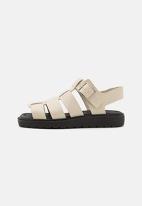 Beige sandalen met meerdere brede banden, een lage zwarte zool en een sluiting met een zijgesp. Duurzaam materiaal met een gladde textuur.