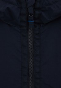 Temno modra jakna z zadrgo dolžine do konca, teksturirano tkanino in kapuco z vrvico za zategovanje. Naluknjani modri šivi vzdolž zadrge za kontrast.