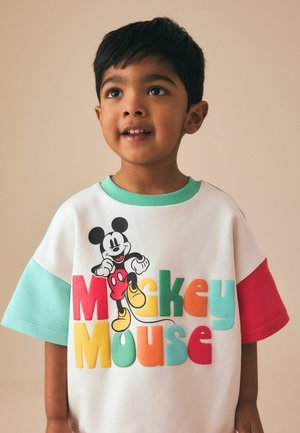 Junger Junge trägt ein buntes Mickey-Mouse-Shirt mit roten, gelben, grünen und türkisfarbenen Ärmeln und schaut leicht nach oben vor einem neutralen Hintergrund.