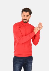 Maglione rosso a collo alto con maniche lunghe e polsini a coste, indossato con jeans blu scuro, tenuto da un modello che aggiusta le maniche.