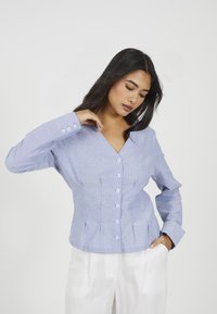 Donna con lunghi capelli scuri indossa una camicetta a righe blu chiaro con bottoni e pantaloni bianchi, pose con una mano vicino al collo e l'altra in tasca.