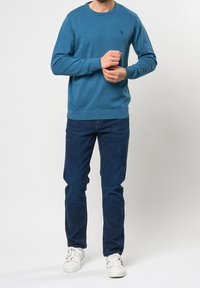 Niebieski dziergany sweter z długimi rękawami, prążkowanym dekoltem i mankietami, z logo na lewej piersi, zestawiony z ciemnymi dżinsami i białymi trampkami.