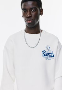 Witte sweatshirt met ronde hals, met een blauwe "Peanuts" graphic en een Snoopy-illustratie op de linkerkant van de borst. Soepele textuur, casual ontwerp.