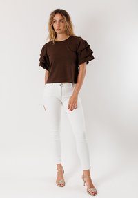 Top marrone a maniche corte con maniche a volant, abbinato a jeans skinny bianchi e tacchi oro con cinturini. Tessuto liscio, vestibilità casual.