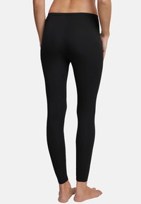 Persona vista desde atrás, vistiendo leggings negros lisos, de pie descalza contra un fondo claro y liso.