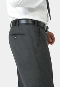 Homme portant un pantalon de costume gris foncé avec une ceinture en cuir noire et une chemise blanche avec une cravate noire, vu de la taille au milieu des cuisses.