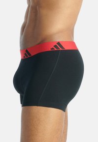 Černé bavlněné boxerky s červeným elastickým pasem, na němž se nacházejí tři černé loga Adidas. Představují přiléhavý střih s detailem šití.