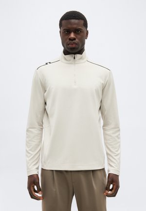 Mann trägt einen hellbeigen Langarm-Halbreißverschluss-Pullover und braune Hose, steht vor einem schlichten weißen Hintergrund.