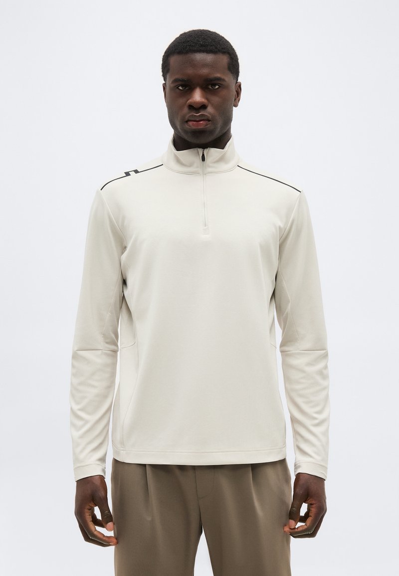 Άνδρας που φοράει ανοιχτό μπεζ μακρυμάνικο πουλόβερ με μισή ζώνη και καφέ παντελόνι, στέκεται μπροστά σε απλό λευκό φόντο.