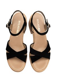 Czarne zamszowe sandały z krzyżującymi się paskami, regulowanym paskiem wokół kostki i niskim drewnianym obcasem. Gładka beżowa wkładka i teksturowana podeszwa.