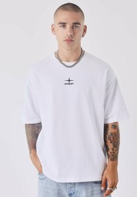 Witte katoenen t-shirt met een zwart logo op de borst. Losse pasvorm, korte mouwen, gecombineerd met denim jeans en zichtbare tatoeages op één arm.
