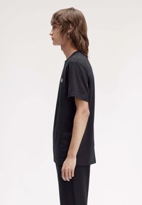 Zwarte katoenen t-shirt met korte mouwen en een ronde halslijn, met een klein geborduurd logo op de borst, getoond op een model van opzij.