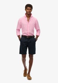 Camicia button-up a maniche lunghe di colore rosa chiaro abbinata a pantaloni corti blu scuro. Cintura di pelle marrone, indossa scarpe in suede beige a taglio basso.