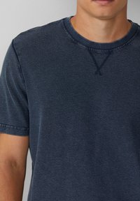 Nahaufnahme einer Person, die ein dunkelblaues, kurzärmliges, strukturiertes T-Shirt mit einem gerippten Rundhalsausschnitt und dekorativen Nähten am Kragen trägt.