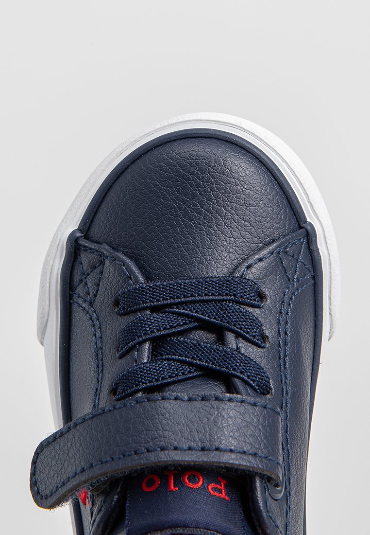 Sneaker en cuir bleu marine avec semelle blanche, présentant un dessus texturé, des lacets et une sangle velcro. Des accents rouges sont visibles sur le côté.