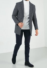 Grijze jas met houndstooth-patroon over een witte coltrui, gecombineerd met donkerblauwe slim-fit broek en zwarte suède enkellaarsjes.