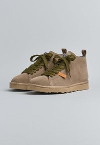Zapatos altos de ante marrón con cordones gruesos en color verde oliva y suelas de goma beige sobre un fondo liso gris claro.