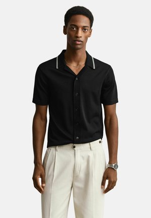 Homme portant une chemise noire à manches courtes avec col bordé de blanc, un pantalon plissé beige, une montre argentée et un fin collier en or.