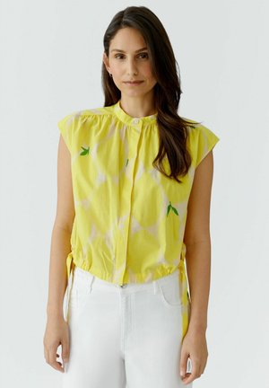 Frau mit langen braunen Haaren, trägt eine ärmellose gelbe Bluse mit grünen Blättern und weiße Hose, steht vor einfachem Hintergrund.