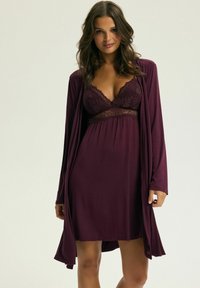 Burgundowa koszula nocna z koronkowymi detalami i dekoltem w serek, noszona pod dopasowanym, otwartym szlafrokiem. Gładki materiał i luźny krój.